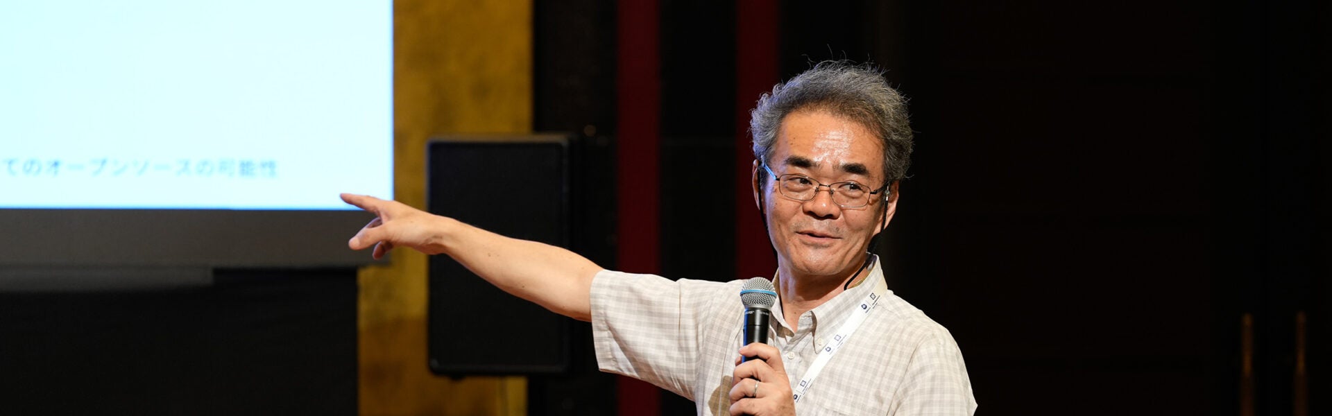A speaker holding a microphone gestures toward a presentation screen during a talk at a conference.