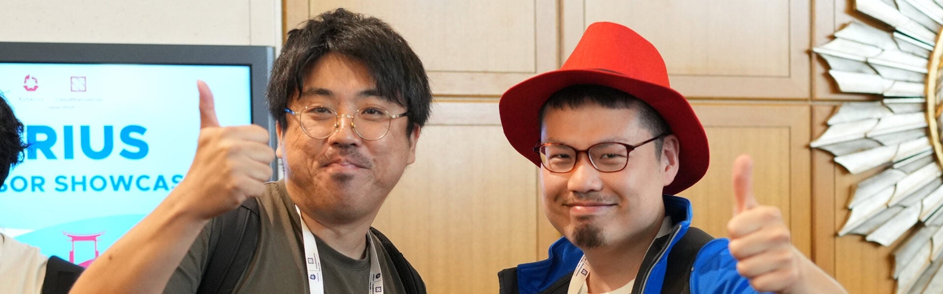 Two people at a conference smiling and giving thumbs-up gestures, one wearing glasses and a red hat, standing in front of event signage.
