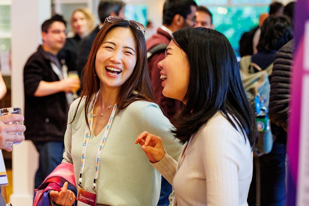 Two conference attendees wearing event badges smile and chat while standing in a busy networking area with other participants in the background.