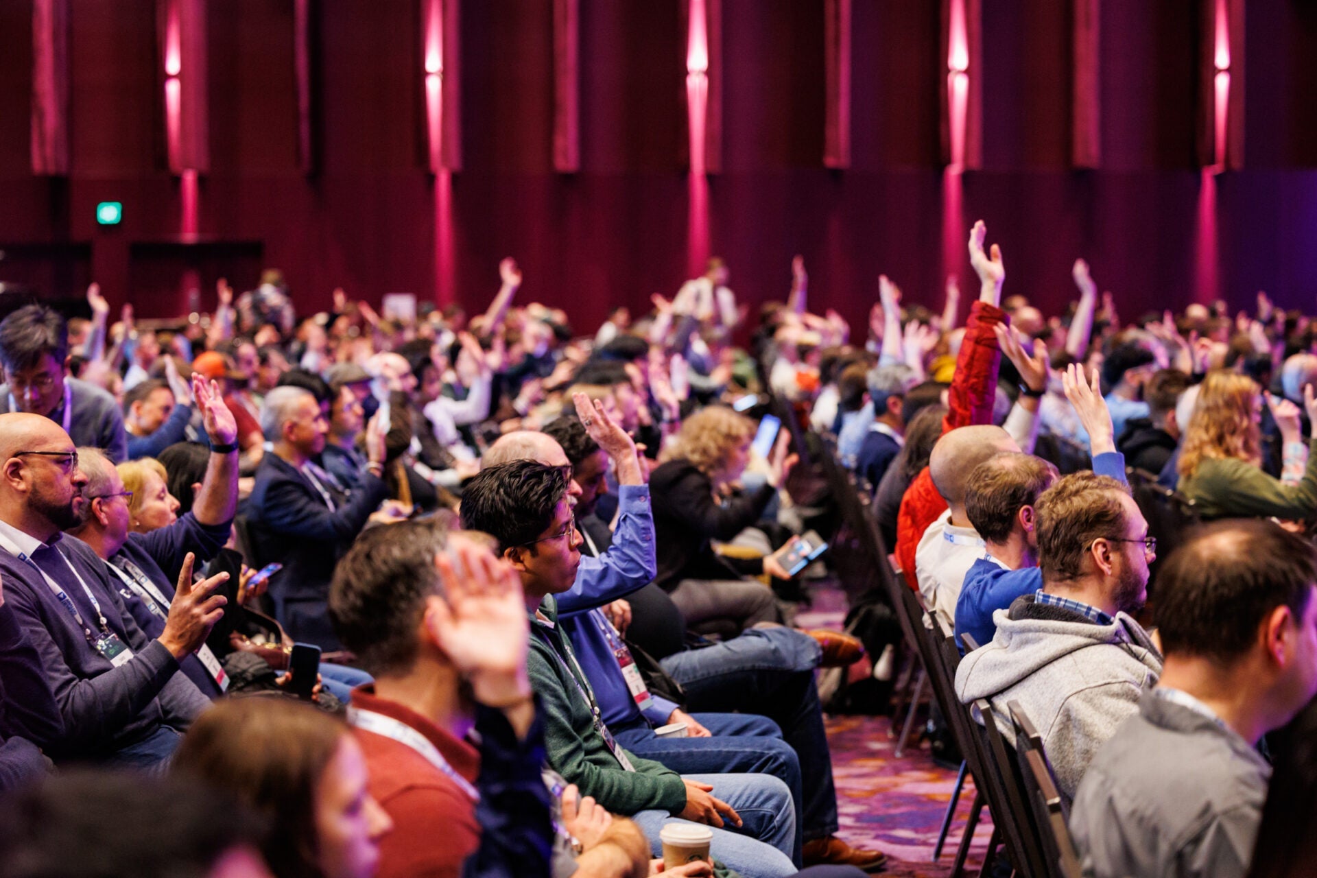 Audience members seated in a conference hall raise their hands during a presentation.