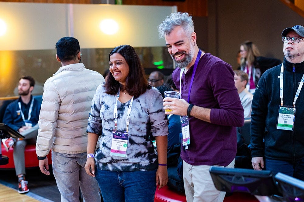 Two attendees wearing conference lanyards laugh together during a lively networking event with a crowd of people behind them.