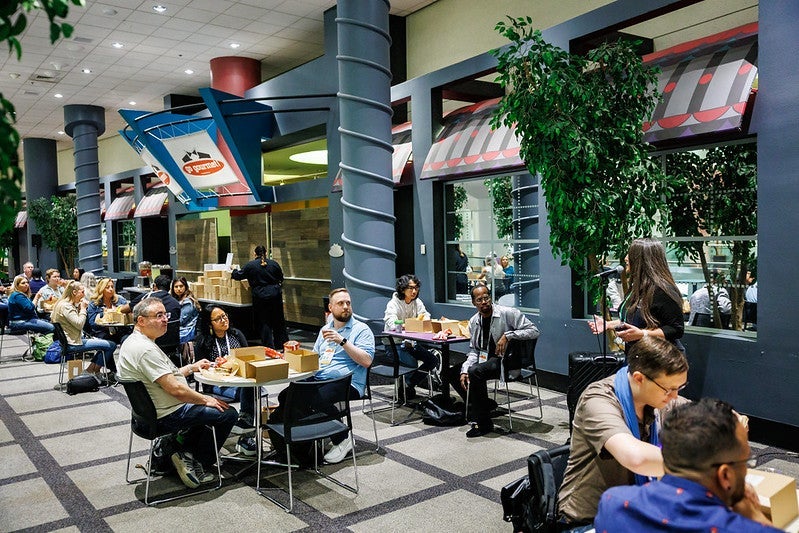 A group of attendees sitting at round tables listening to a speaker at a microphone.