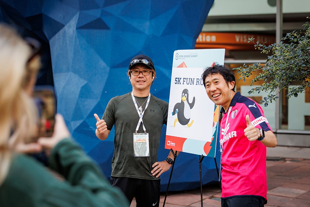 Two attendees pose with thumbs up beside a “5K Fun Run” sign during a conference outdoor activity.