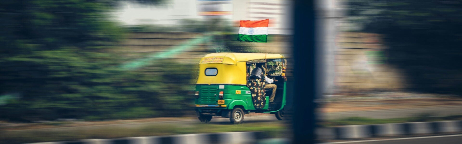 Green and yellow auto rickshaw driving past with motion blur, with an Indian flag visible in the background