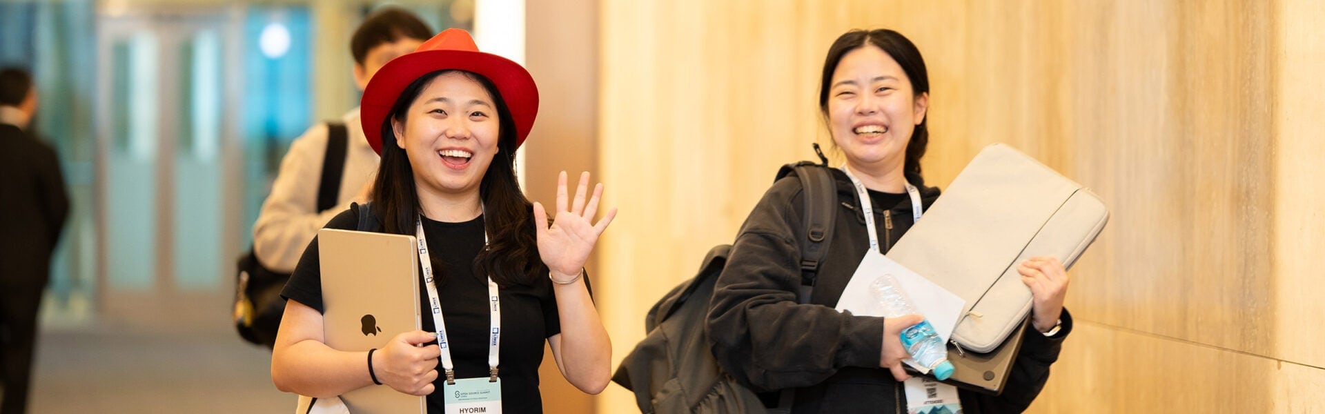 Two conference attendees wearing event badges walk down a hallway, smiling as one waves while holding a laptop.