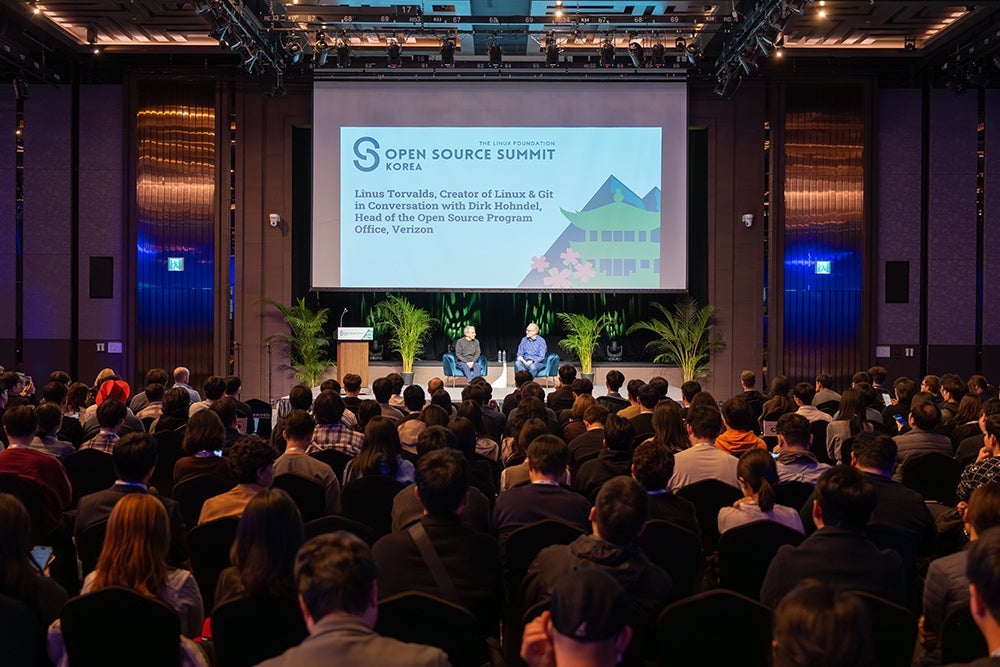 Wide view of a large conference hall during Open Source Summit Korea, with two speakers seated on stage beneath a large presentation screen while a full audience listens.