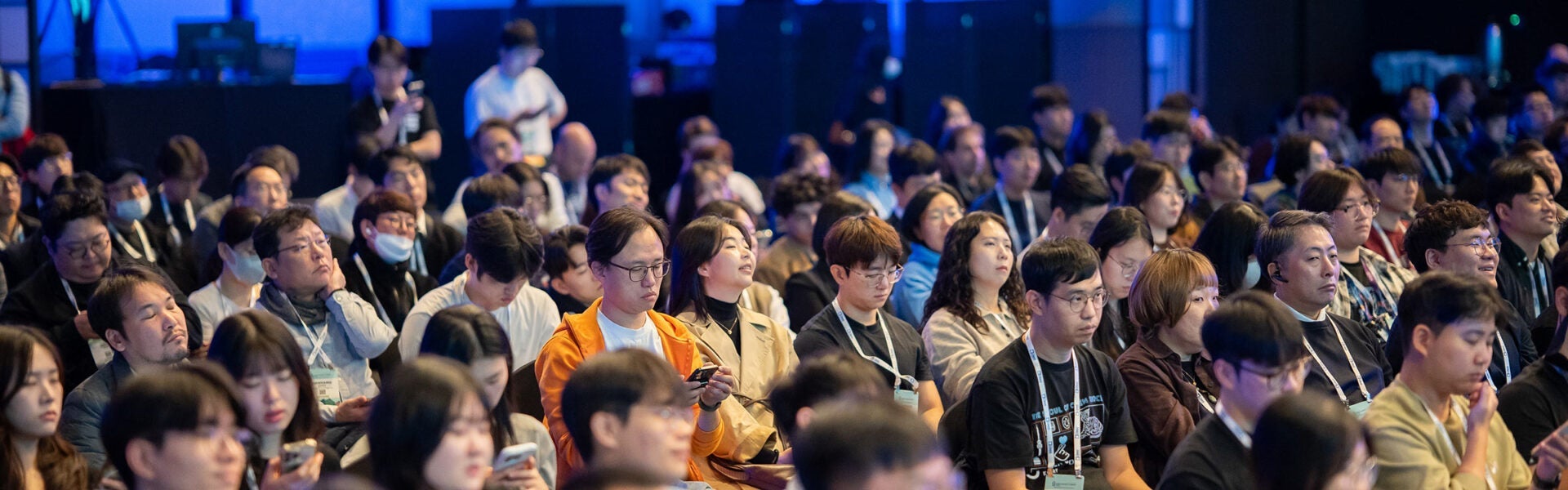 Audience members seated closely together during a conference session, many wearing badges and looking toward the stage while some check their phones.