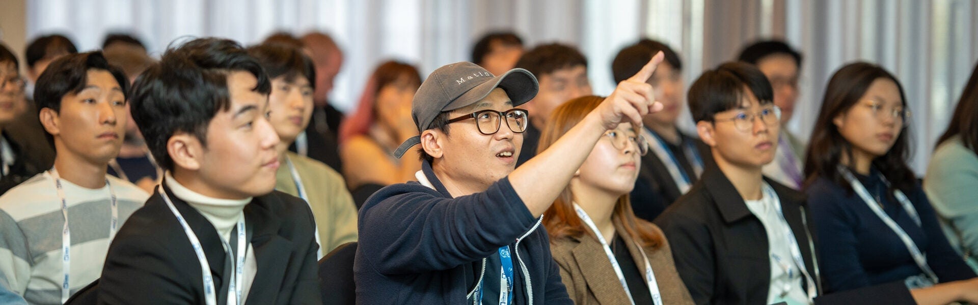 Attendee in a cap raises a hand and points during a conference session, surrounded by other seated participants wearing event badges.