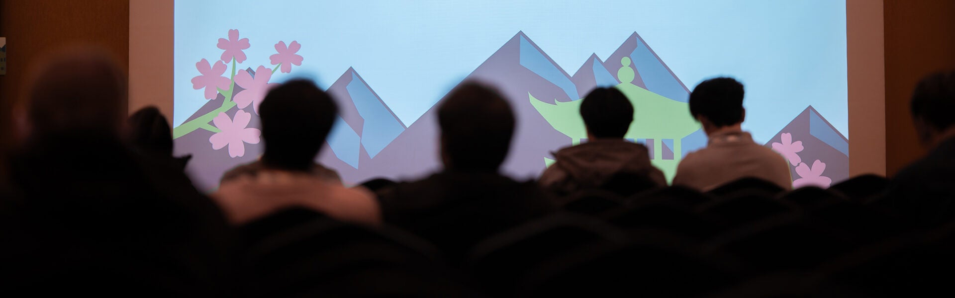 Audience members seated in a darkened conference room watching a presentation projected on a screen with stylized mountains and cherry blossoms.