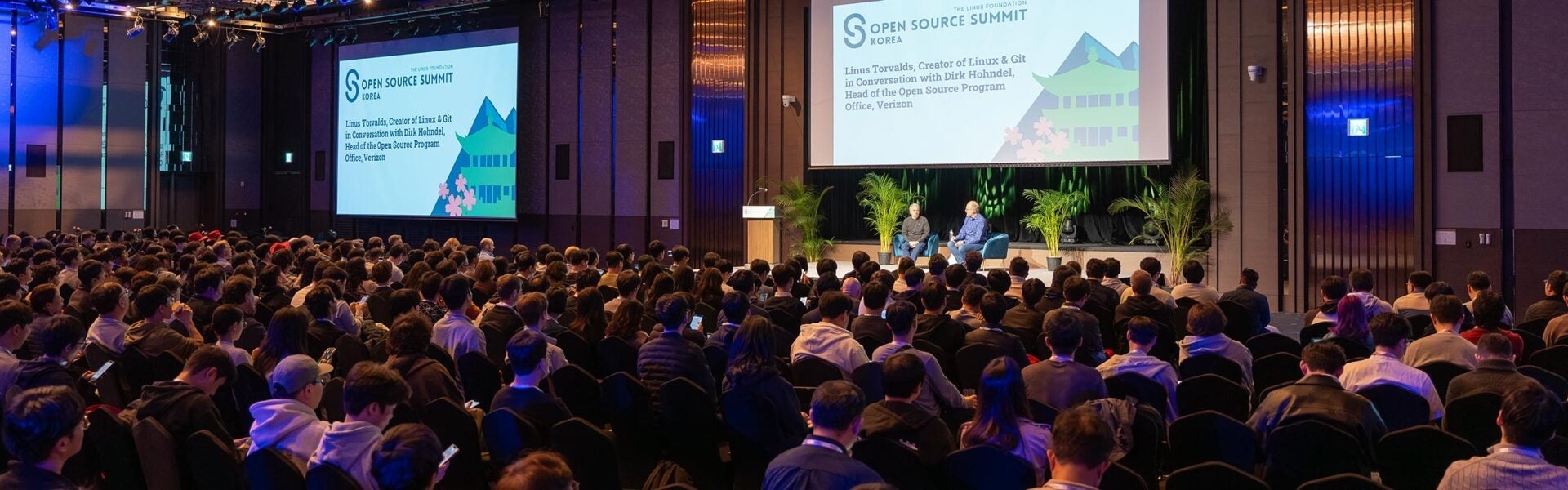 Large audience seated in a conference hall watching a stage discussion at Open Source Summit Korea, with presentation screens visible behind the speakers.