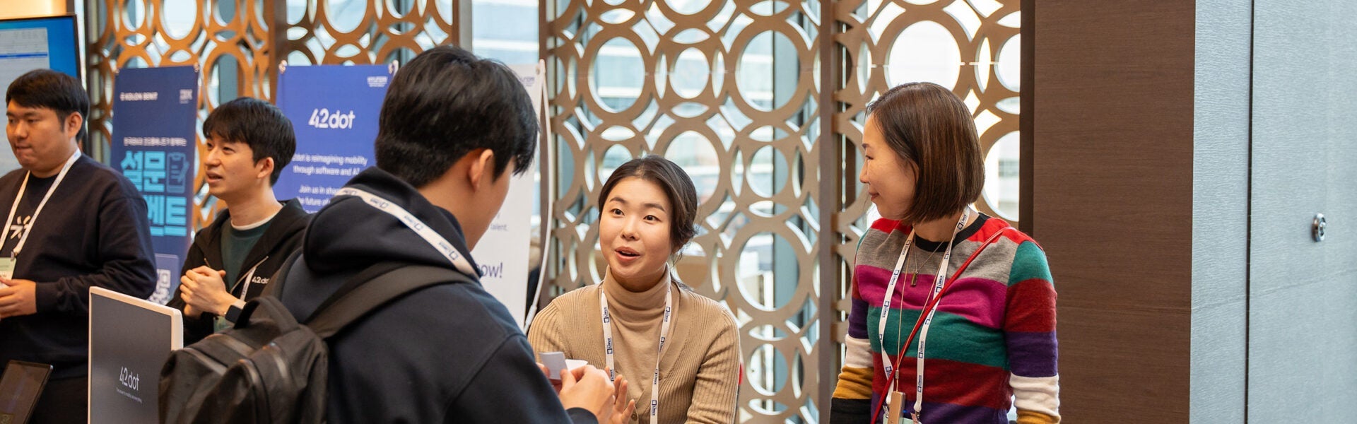 Conference attendees speaking with representatives at a sponsor booth area, with informational displays and event badges visible.