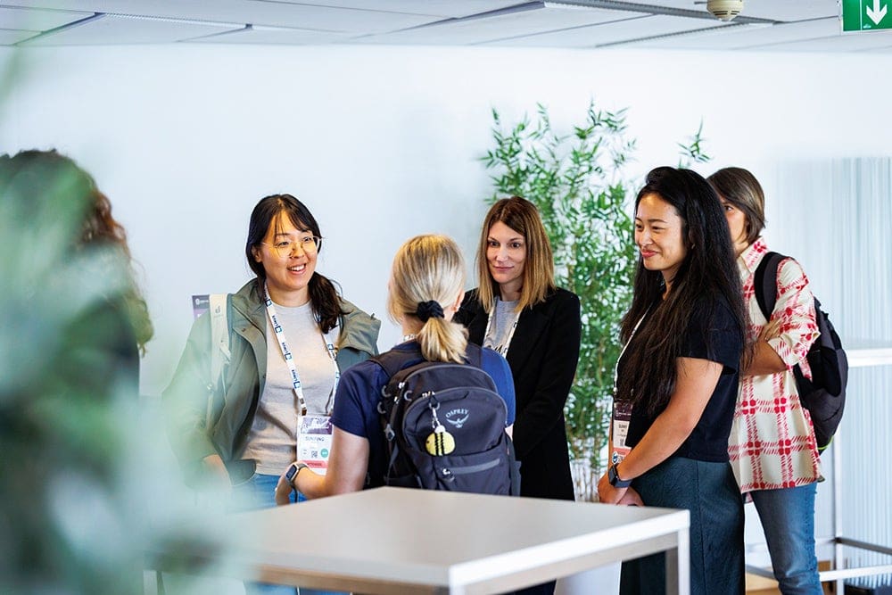 Five conference attendees stand together in a bright networking area, talking casually during an event.