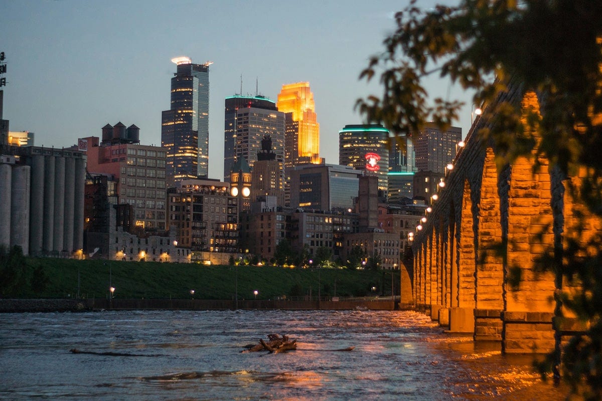 Downtown buildings in the background of water and a bridge lit up at dusk.
