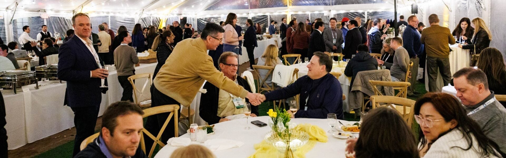 Conference attendees seated at round tables while one attendee greets another with a handshake in a large event space.