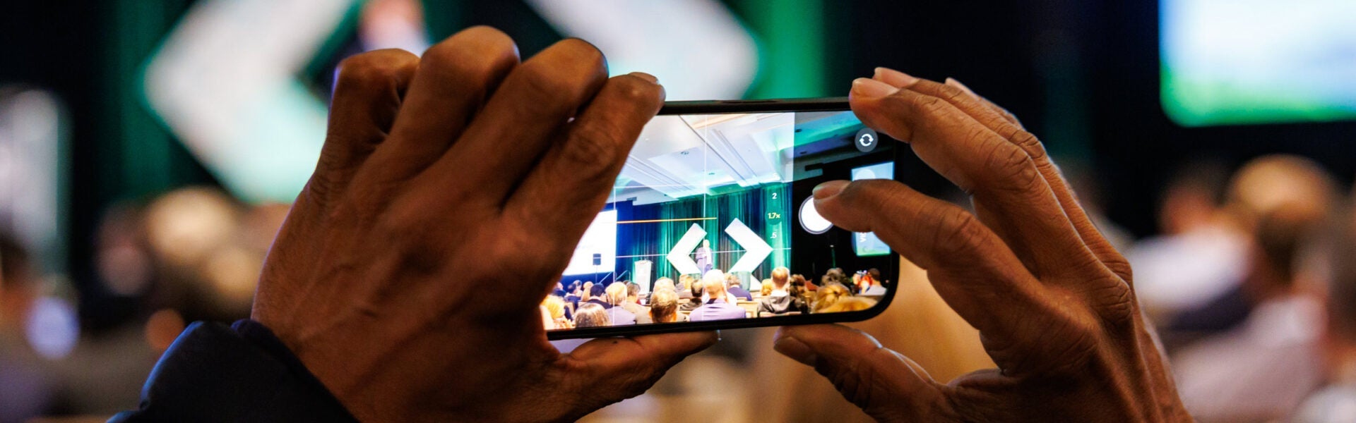 Attendee holding a smartphone to photograph a speaker on stage during a conference session.