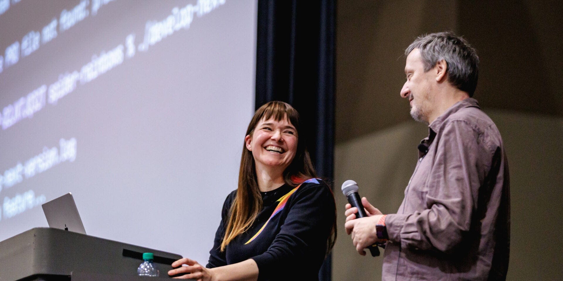 Two speakers smiling on stage during a presentation, with one holding a microphone.