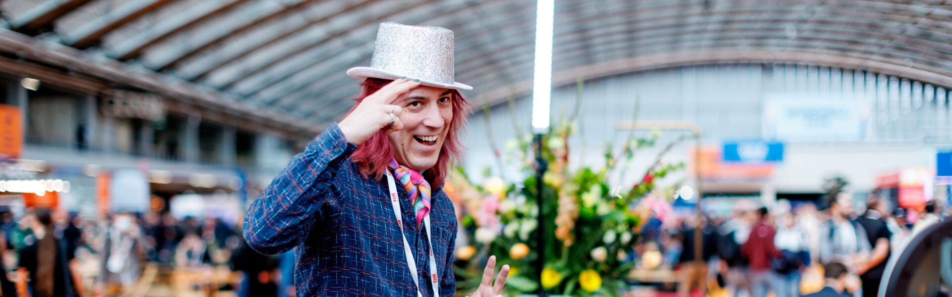 Person with red hair wearing a silver top hat and colorful scarf, smiling and saluting the camera at a busy conference venue.