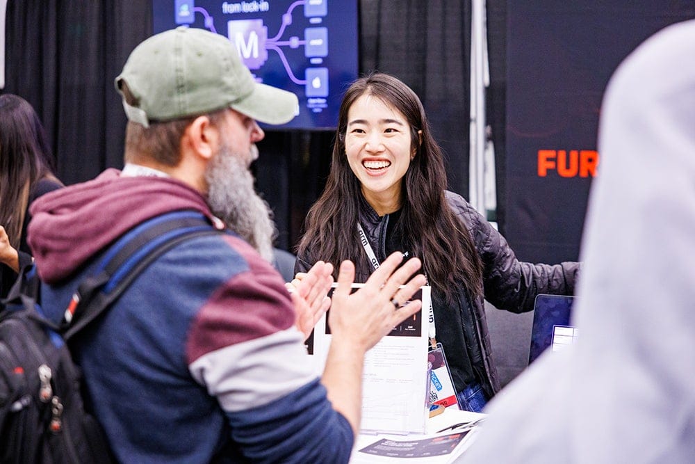 Conference attendee smiling and talking with another attendee at an exhibitor booth.